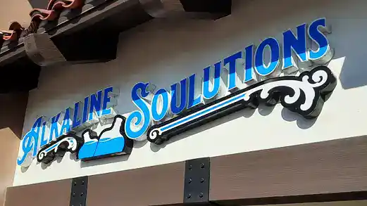 Custom storefront sign for Alkaline Solutions featuring illuminated cursive and bold lettering with a blue water-inspired design, installed on a tiled-roof building in Vista, CA.