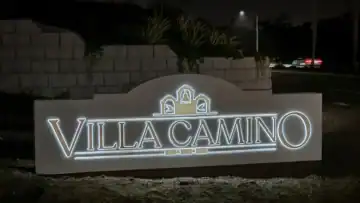Illuminated “Villa Camino” monument sign at night, featuring three decorative arches with bell motifs, set against a landscaped stone wall and street backdrop in Oceanside, California. The sign is solar powered.