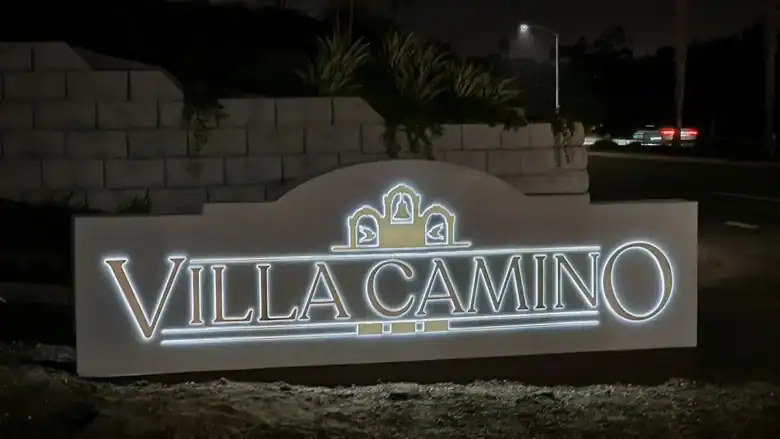 Illuminated “Villa Camino” monument sign at night, featuring three decorative arches with bell motifs, set against a landscaped stone wall and street backdrop in Oceanside, California. The sign is solar powered.