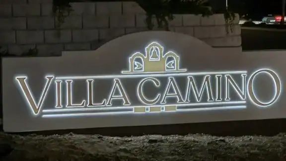 Illuminated “Villa Camino” monument sign at night, featuring three decorative arches with bell motifs, set against a landscaped stone wall and street backdrop in Oceanside, California. The sign is solar powered.