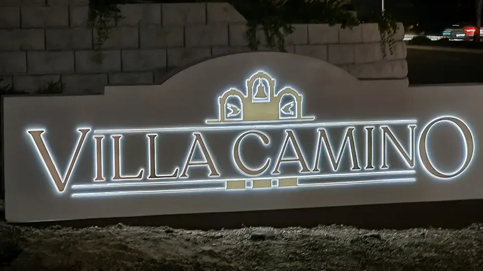 Illuminated “Villa Camino” monument sign at night, featuring three decorative arches with bell motifs, set against a landscaped stone wall and street backdrop in Oceanside, California. The sign is solar powered.