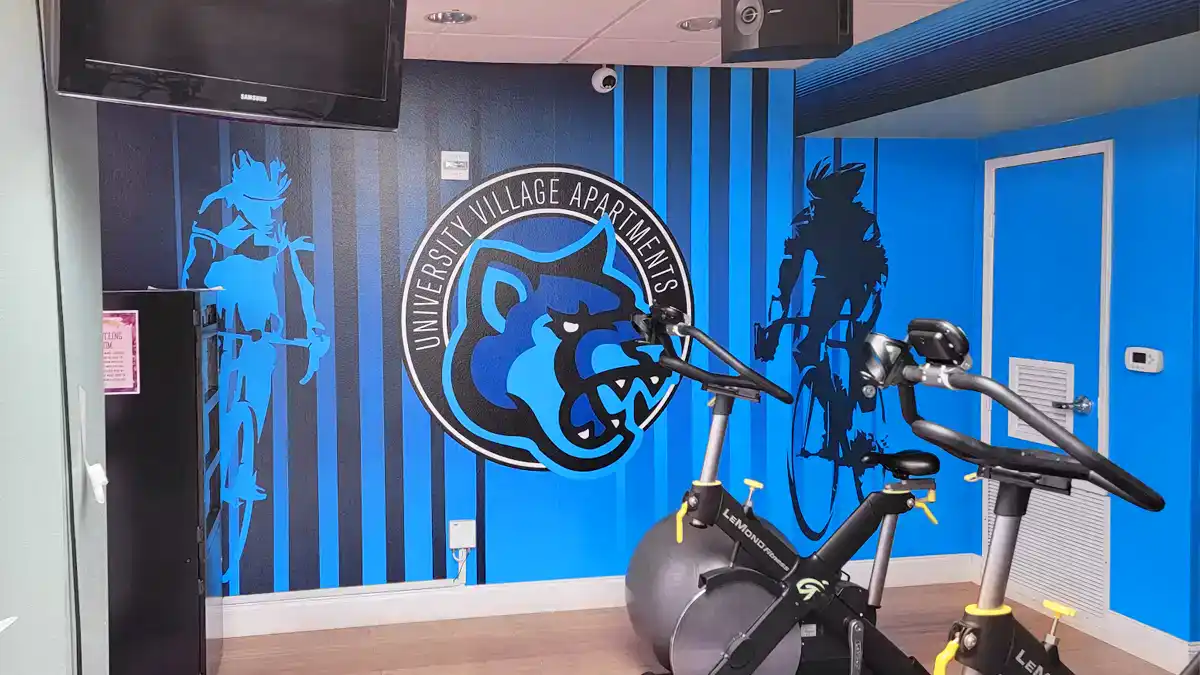 Custom wall graphics installed in the cycle room of University Village Apartments in San Marcos, featuring the blue Cougar mascot logo and silhouettes of cyclists.