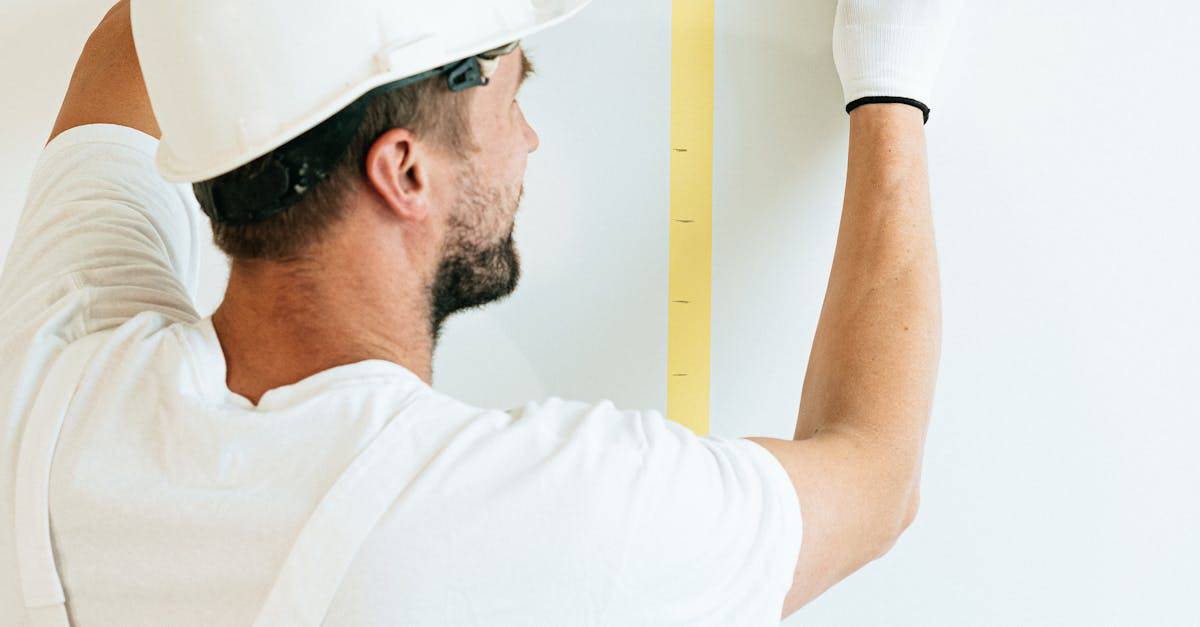 A man in a hard hat and gloves measuring a wall with a tape measure.
